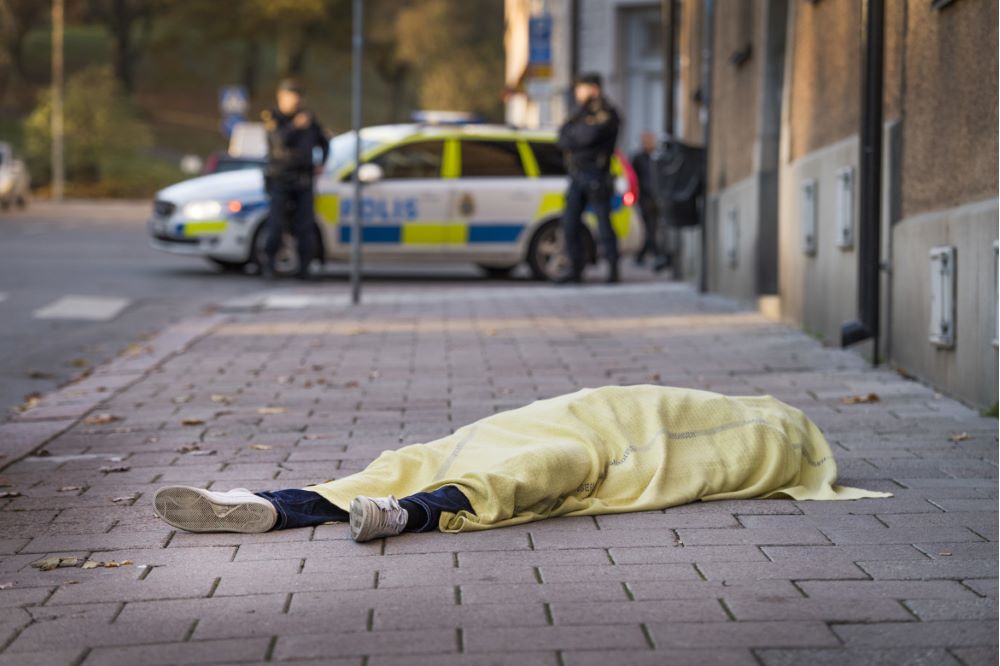 Död person som ligger på trottoar. Poliser och en polisbil syns i bakgrunden.