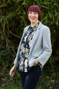 This is a picture of Lisa. Lisa is a white woman with short, red hair. She has a fringe, and is smiling. She is standing, with one hand in the pocket of her trousers. She is wearing a flowery shirt, a pair of dark trousers, and a grey blouse. She is looking straight into the camera.