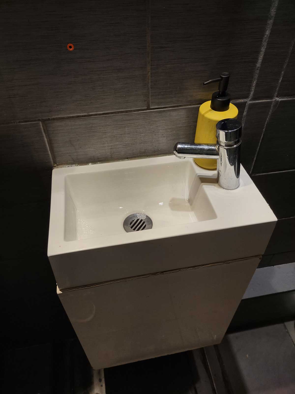 Compacte witte keramische wastafel met een chromen kraan staat tegen een donkere tegelwand. Een felgele zeepdispenser (10-5-2026)