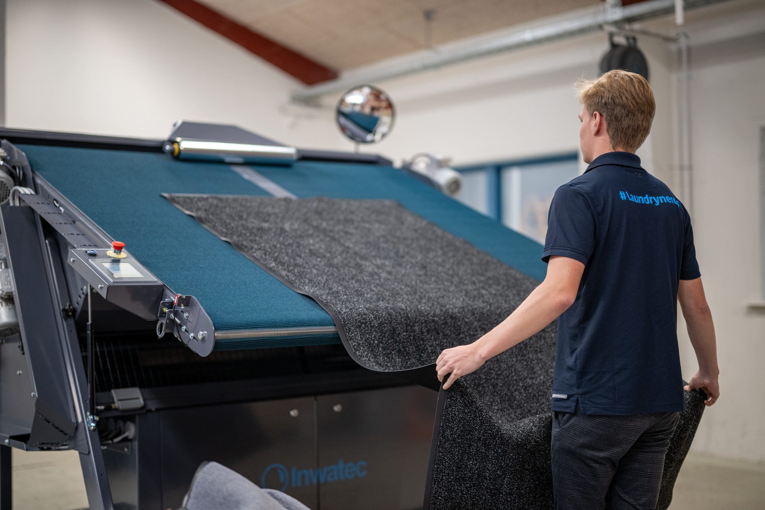 Inwatec employee with #laundrynerds on the back is showing how to feed mats into the Typhoon Mat Roller machine.