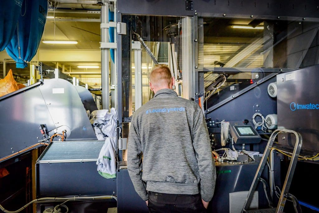Inwatec employee standing in front of a Inwatec machine. On the back of the employees shirt you see the text #laundrynerds.