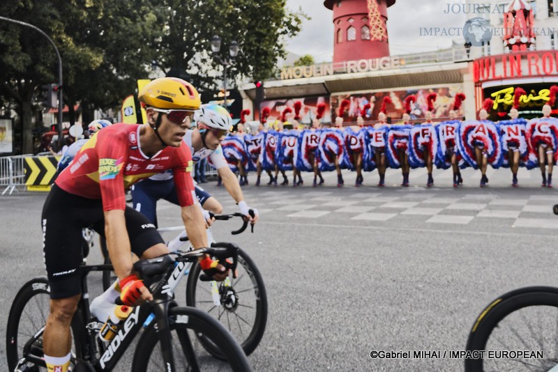 Plusieurs coureurs du Tour de France passent devant le Moulin Rouge tandis que les danseuses poursuivent leur spectacle place Blanche à Paris.