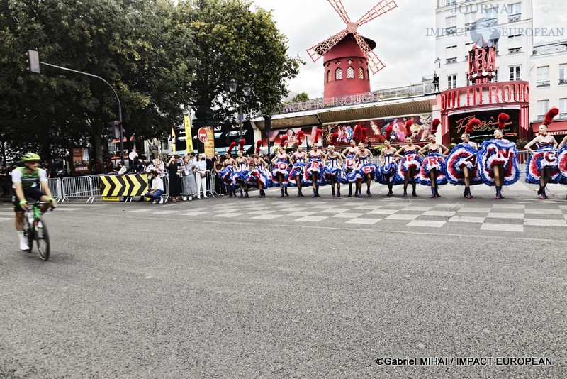Un coureur du Tour de France quitte la place Blanche tandis que les danseuses du Moulin Rouge poursuivent leur spectacle devant le célèbre cabaret parisien.