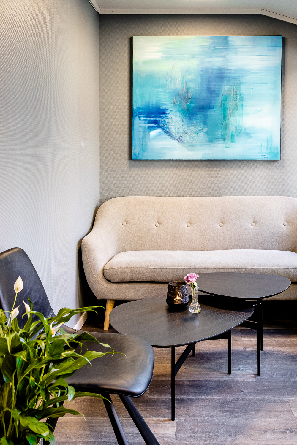 Interiørbilde av sofakroken hos Kafé Glede. Over sofaen henger et flott maleri som går i blåtoner. Foran sofaen er det satt opp to små bord og en ekstra stol. Nærmest står en plante.