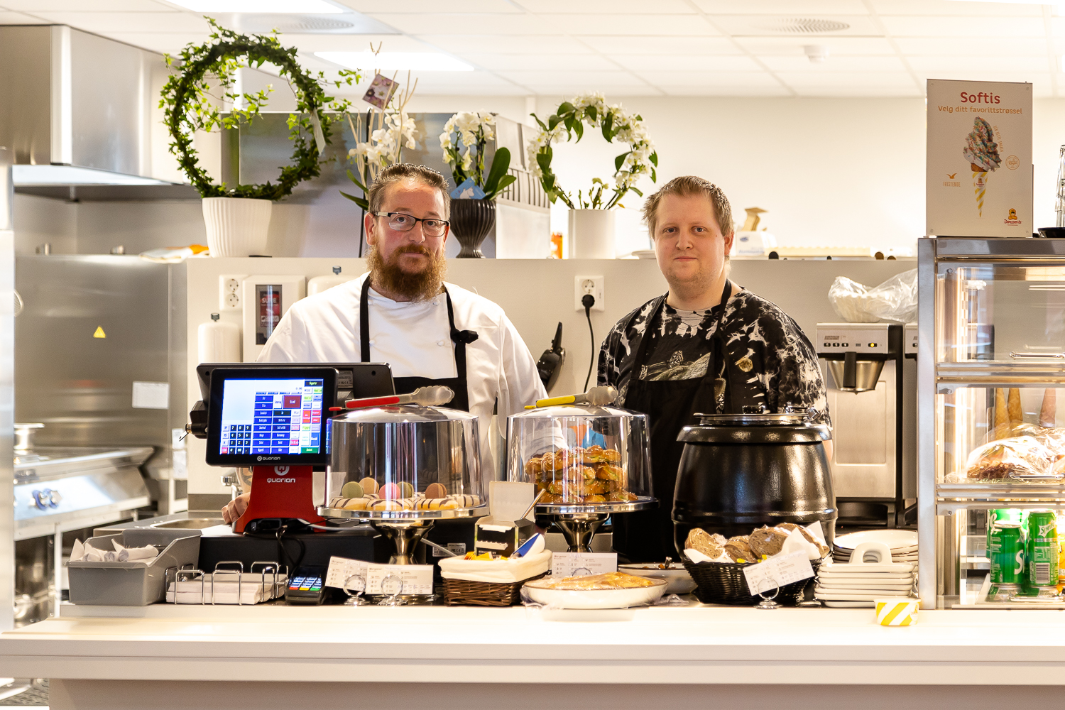 Nærbilde av to medarbeidere fra Kafé Glede. De står bak disken der de tilbyr makroner og små kaker, i tillegg til dagens suppe. På hylla bak står fire buketter kafeen fikk på åpningsdagen i november 2021. Vi skimter såvidt kjøkkenet i bakgrunnen og ser også en skilt som markedsfører at de lager softis.