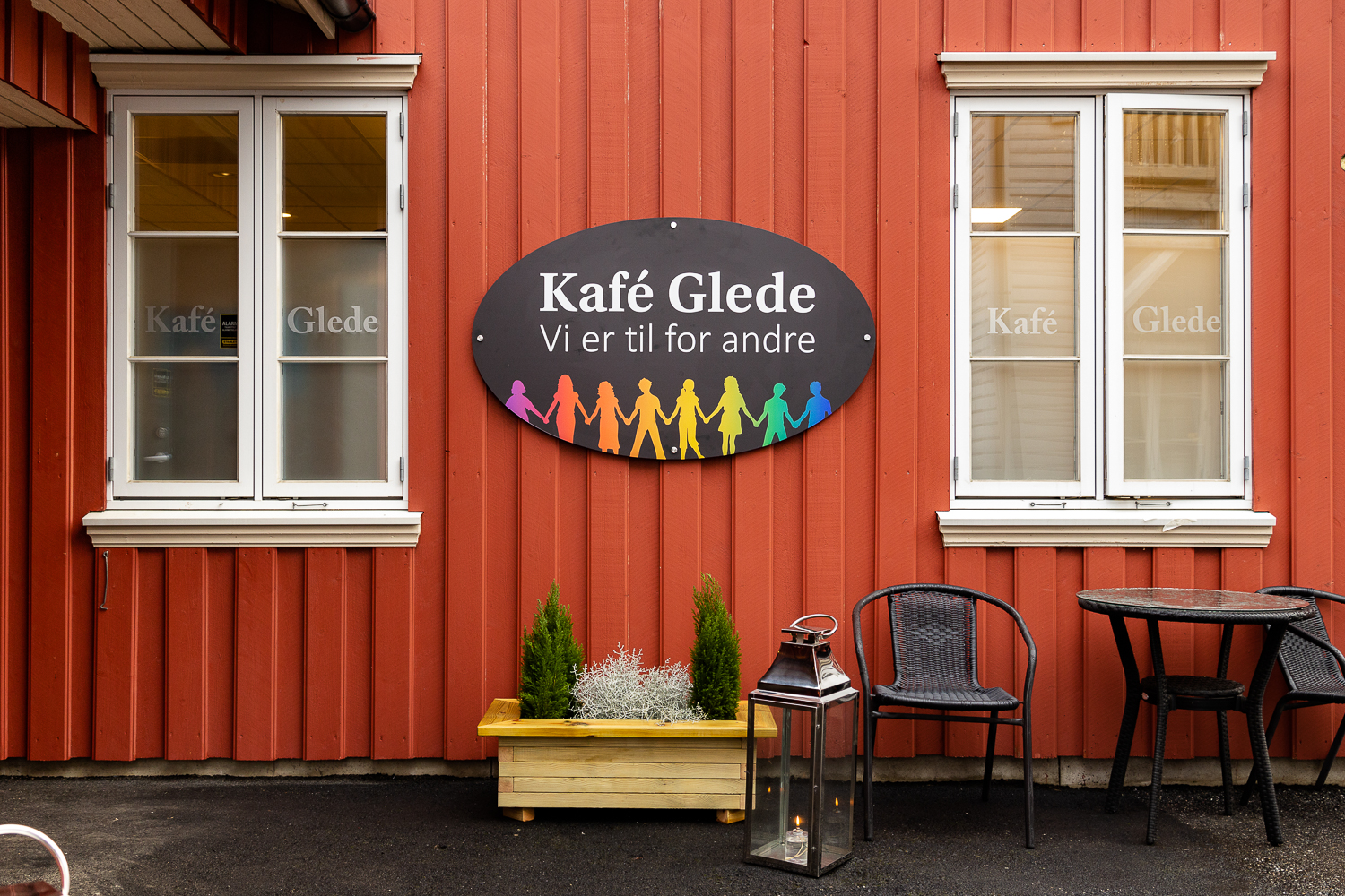 Bilde tatt utenfor hovedinngangen til Kafé Glede. Midt i bildet er logoen til kafeen, der bedriftens navn er nevnt i tillegg til slagordet "Vi er til for andre". Nederst på logoen er den regnbuefargede menneskelenken. Skiltet er hengt opp på en rød panelvegg med to vinduer. Det er arrangert en sitteplass til høyre i bildet.