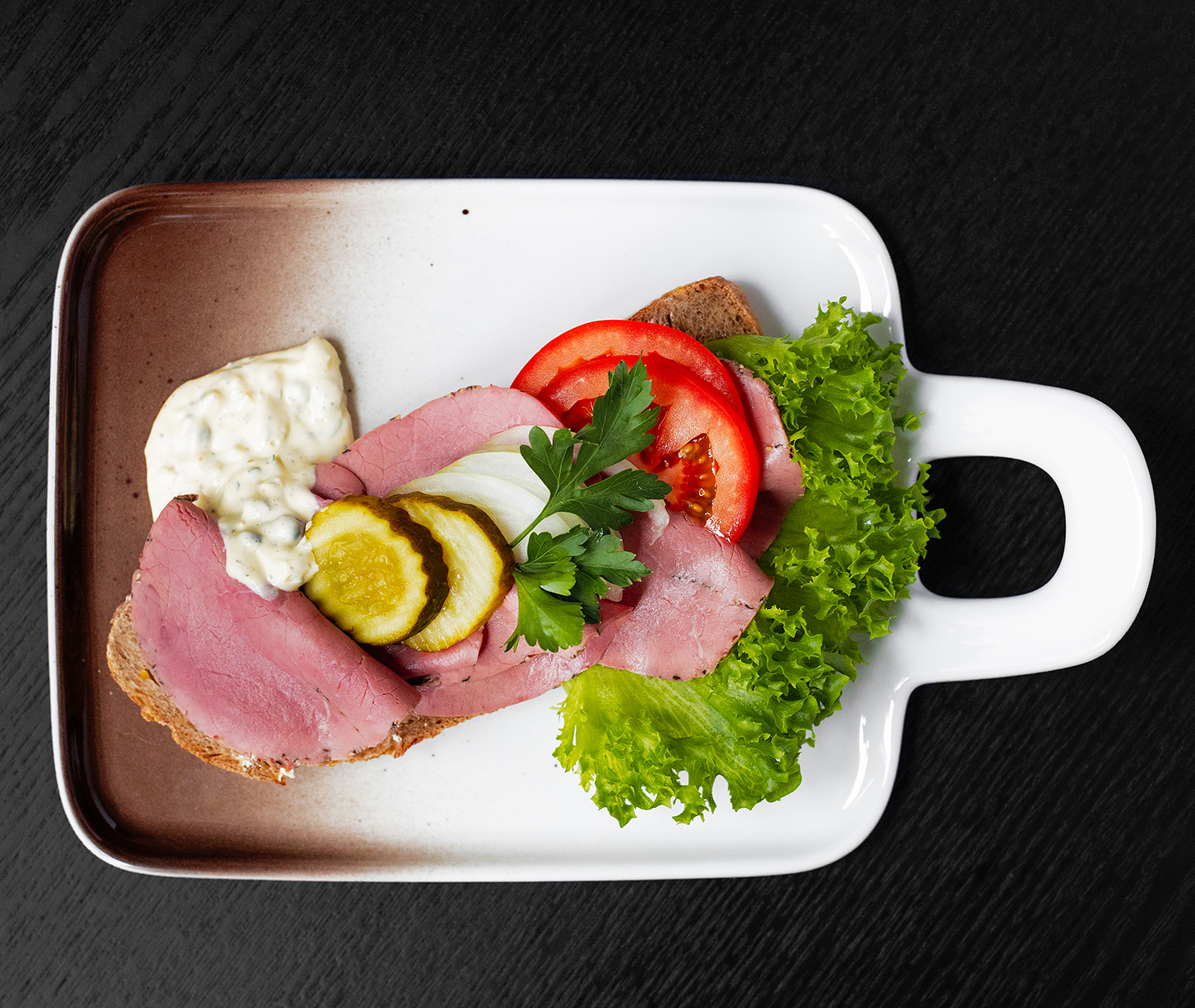 Bilde av smørbrød med roastbiff med remoulade. Oppå roasbiffen er det sylteagurk, løk, tomat, salat og en urt. Maten sees ovenfra liggende på et flott designerfat med håndtak plassert på svart bakgrunn.