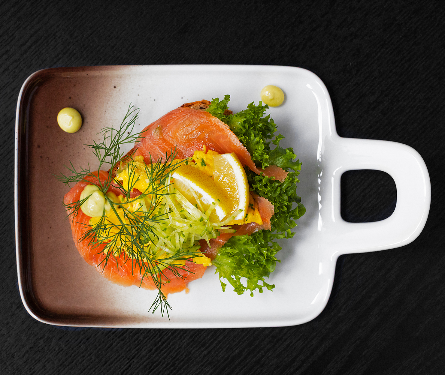 Bilde av smørbrød med laks, sitron, strimlet agurk, dill, eggerøre og salat. I tillegg er det dekorert med noen punkter majones. Maten sees ovenfra liggende på et flott designerfat med håndtak plassert på svart bakgrunn.