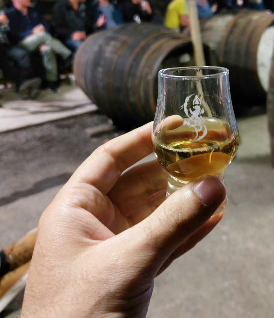 Zu sehen ist die Probe des 16jährigen Lagavulin im Glencairn-Glas, mit zwei Fässern im Warehouse im Hintergrund.