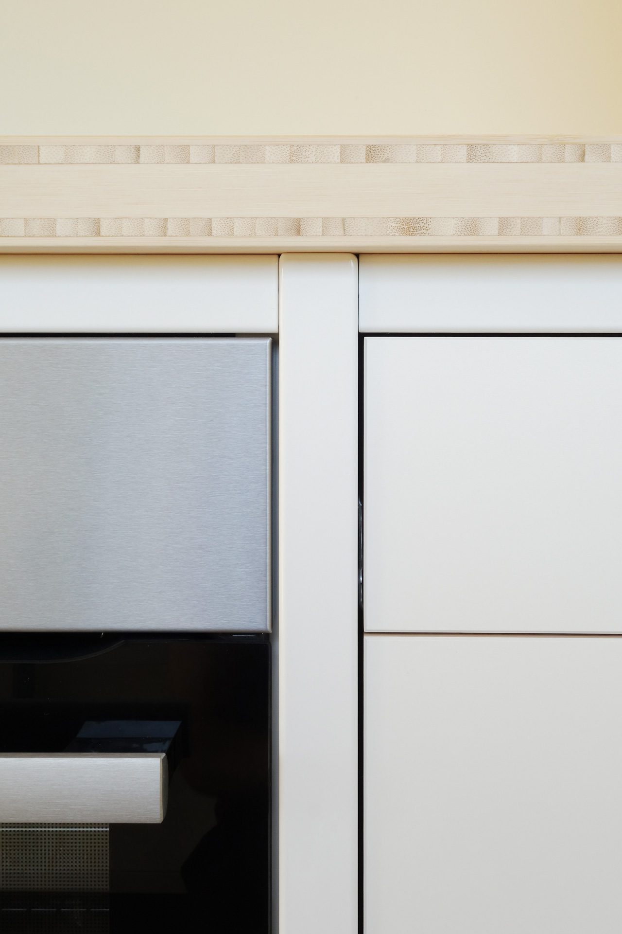 detail-bamboe-werkblad-40mm-keukenfront-wit Detailopname van de witte stalen keukenconstructie tussen een roestvrijstalen oven en witte kastfronten met een bamboe werkblad