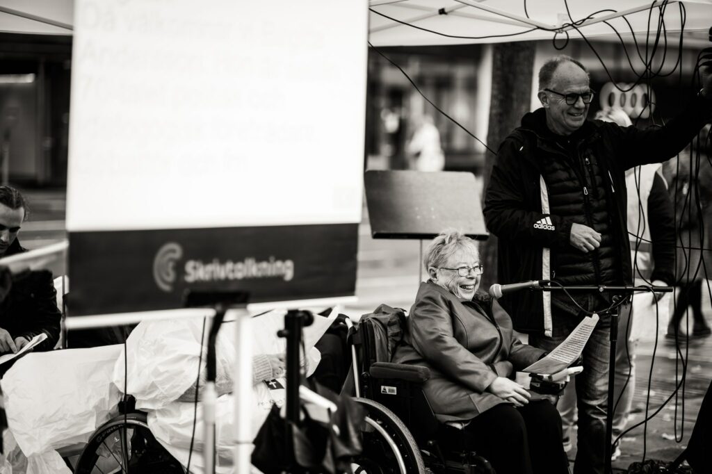 Svartvitt foto. Birgitta Andersson sitter vid mikrofon, Anders ordnar med ljudet bredvid, skärm för syntolkning. Birgitta och Anders skrattar.