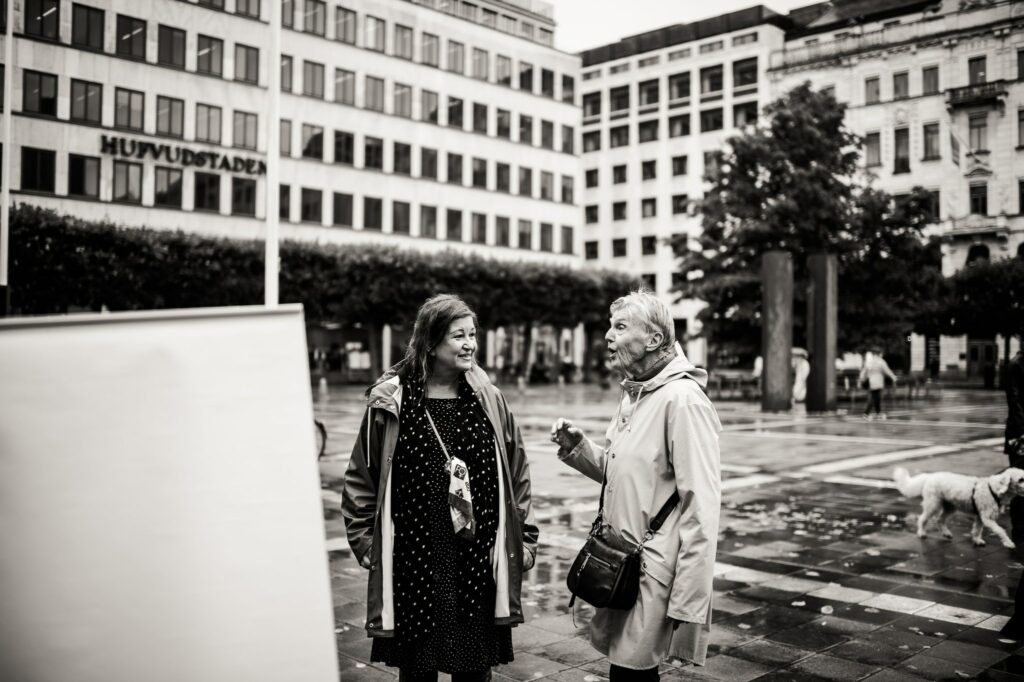 Svartvitt foto. Anne från styrelsen i samtal med Barbro Westerholm på regnigt torg.