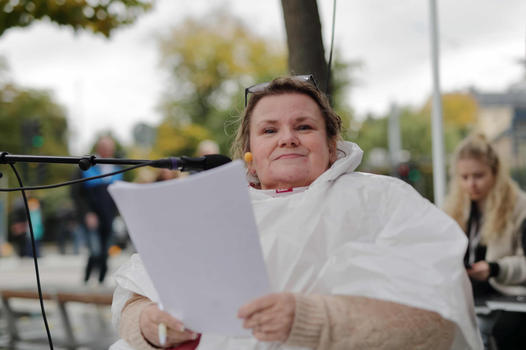 Maria har en vit regncape, i halsen skymtar en FQ-scarf och uppskjutet i pannan sitter glasögon med svarta bågar. Hon sitter ned och har en mikrofon på stativ, samt en ”mygga”. Bakom henne skymtar träd samt människor som rör sig längs trottoaren bakom.