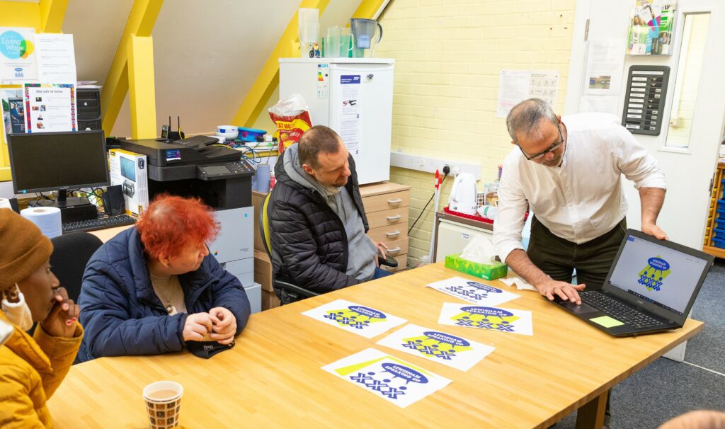 three self advocates - two female, one male - sitting at a table viewing something on a laptop, which is being pointed to by Marsh, a member of the LSU team, standing up. There are print outs on the table