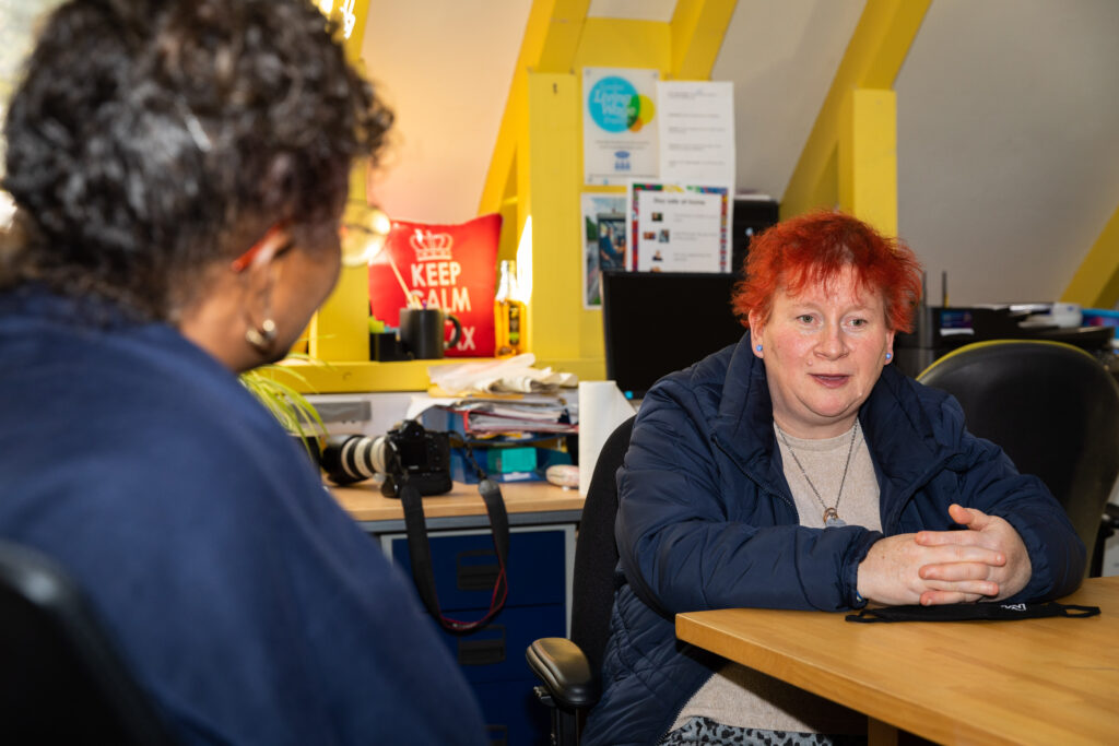 woman with red hair sitting at a table with her arms resting on the table; she is talking to a female advocate whose back is to the camera