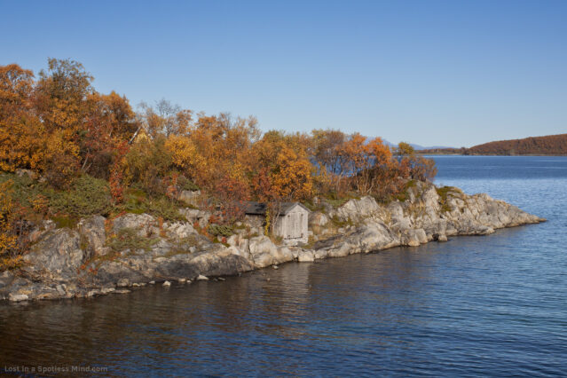 A photo of the seaside, with a rocky shore, autumnal trees, and a bright blue sky and ocean.