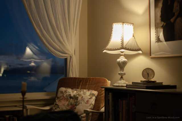 A corner of a room with an ornate lamp and a velvet chair, the golden light inside contrasting against the blue outside the window. A clock shows 5:30 pm.