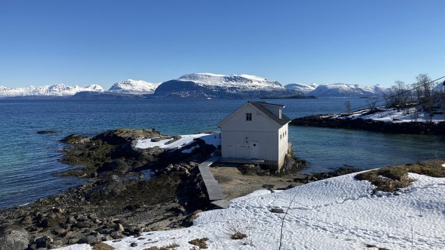 Photo of a seascape, taken in bright sunlight, with snowy mountains, clear blue water, and a white boathouse.