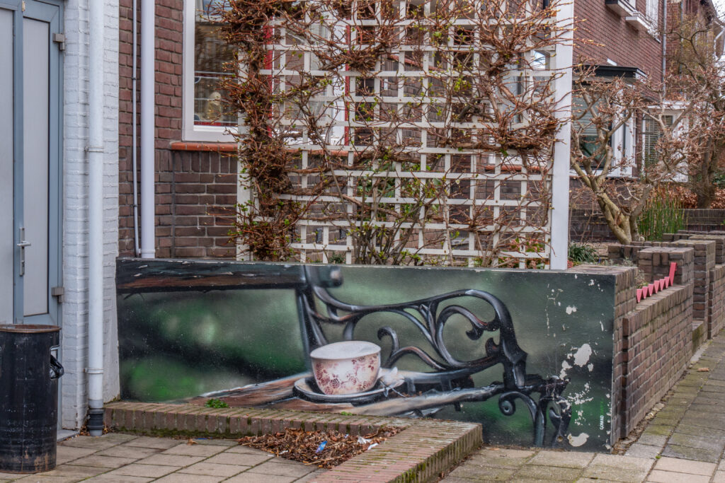 Deze mooie en gezellige muurschildering van een bankje met daarop een kopje koffie prijkt op een klein muurtje tegenover de supermarkt in het hart van Oud-Geleen. Deze muurschildering nodigt uit tot een praatje en misschien zelfs wel tot een kopje koffie.