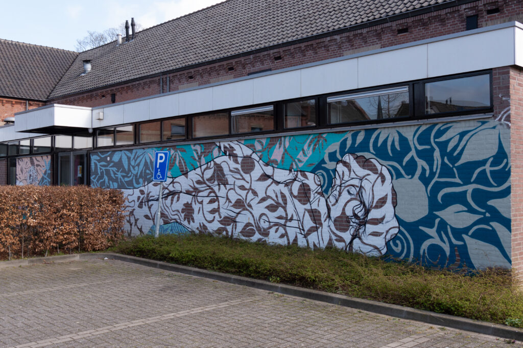 Gijs Scholten heeft vooraf geen ontwerp en dat is ook niet zijn manier van werken. De vorm van de muur en de omgeving bepalen het beeld.
In de afbeelding zie je natuurlijke vormen, een patroon van bloemen en natuur. Voor hem een natuurlijke keuze omdat hij vindt dat dit aansluit bij Lichtenvoorde.
De liggende vrouw is spontaan ontstaan. De vrouw is op afstand verweven met het organische patroon op de achtergrond en lijkt er in op te gaan. Dichterbij zie je de details. De schetsmatige en losse lijnen bieden slechts hulp aan de kijker en zijn in deze voorstelling dan ook niet bepalend, eerder prikkelend.