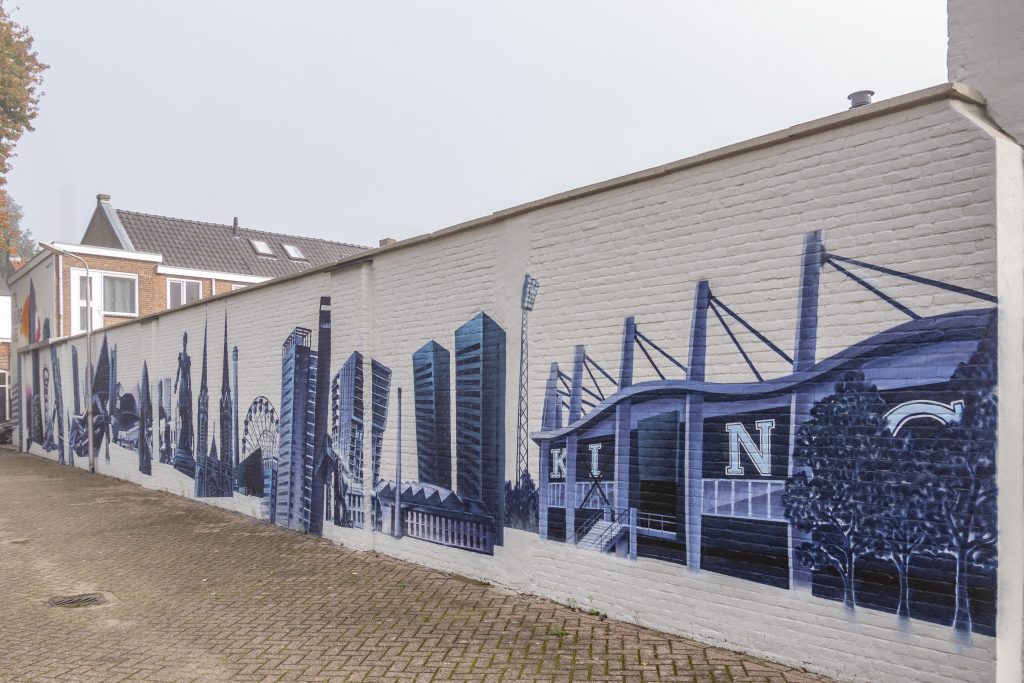 De muur toont de skyline van Tilburg. Er staan verschillende taferelen zoals het reuzenrad van de kermis, de Beka fabriek, het Willem II stadion en de uitkijktoren in het Spoorpark