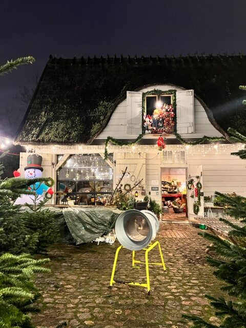 Et billede af julemarked på Stalden på Frederiksberg. Udenfor kan man købe juletræer, indenfor julepynt og julegaver. Det er aften på billede, og der er en masse julelys.