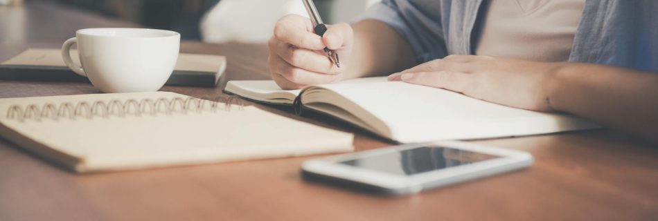 woman writing on a notebook beside teacup and tablet computer