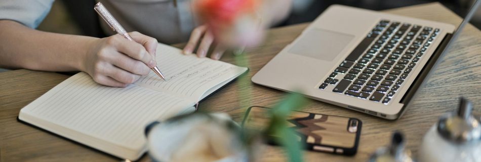 person writing on a notebook beside macbook