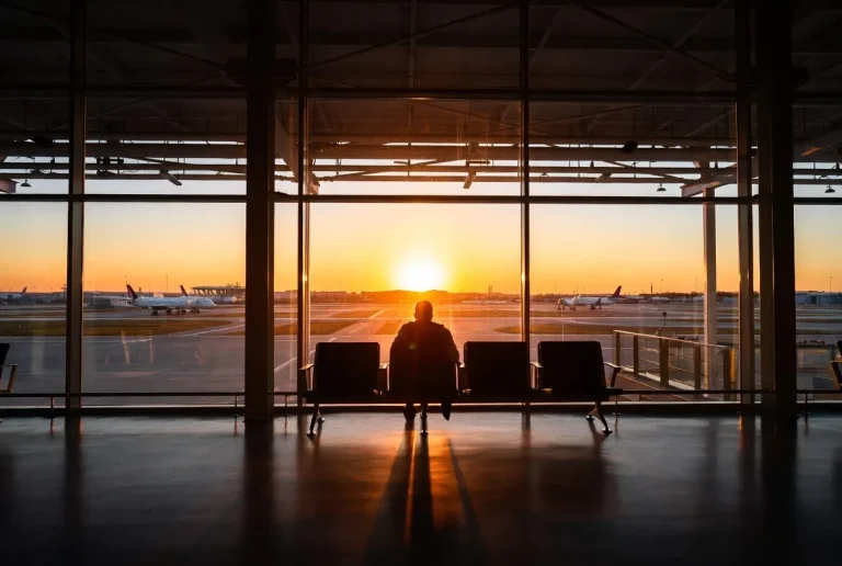 Solo traveler's picture. A lone male figure silhouetted against a vibrant sunset, viewed from an airport terminal window. The tarmac glows as planes await their journeys.
