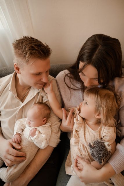 En nyföddfotografering med en familj som sitter i soffan, pappan håller den nyfödda bebisen och mamman håller deras treåriga dotter, som i sin tur klappar på pappans kind.