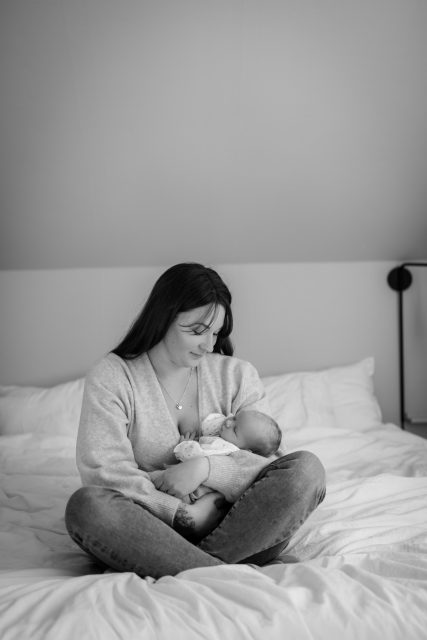 Nybliven mamma sitter på en säng och håller sin nyfödda bebis på nyföddfotografering i Stockholm