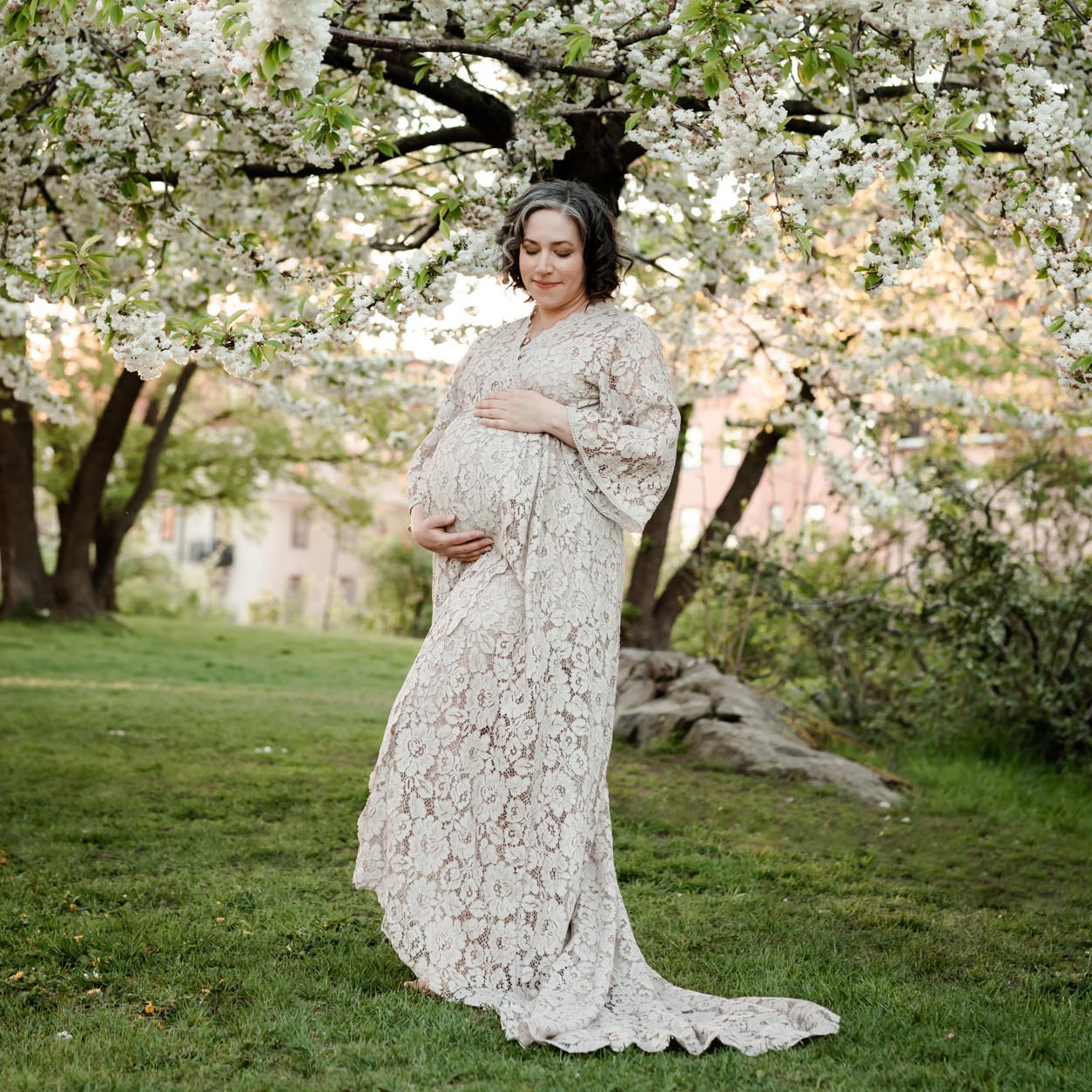 Gravid kvinna i spetsklänning står under ett blommande äppelträd i Stockholm.
