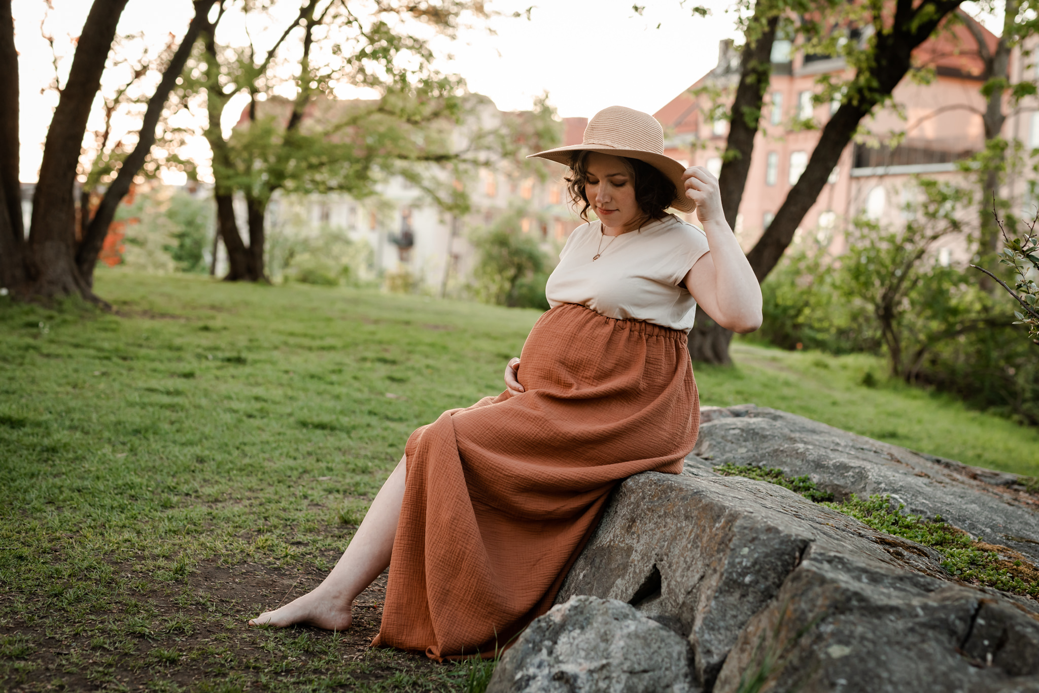 Gravid kvinna sitter på en sten i en park iklädd rostbrun kjol, beige t-shirt och beige hatt