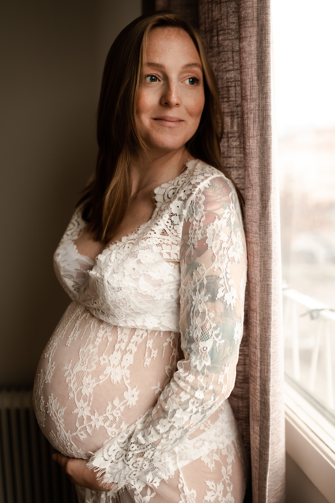 Gravid leende kvinna står med ryggen mot ett fönster iklädd en vit spetsklänning, blickandes ut genom fönstret.
