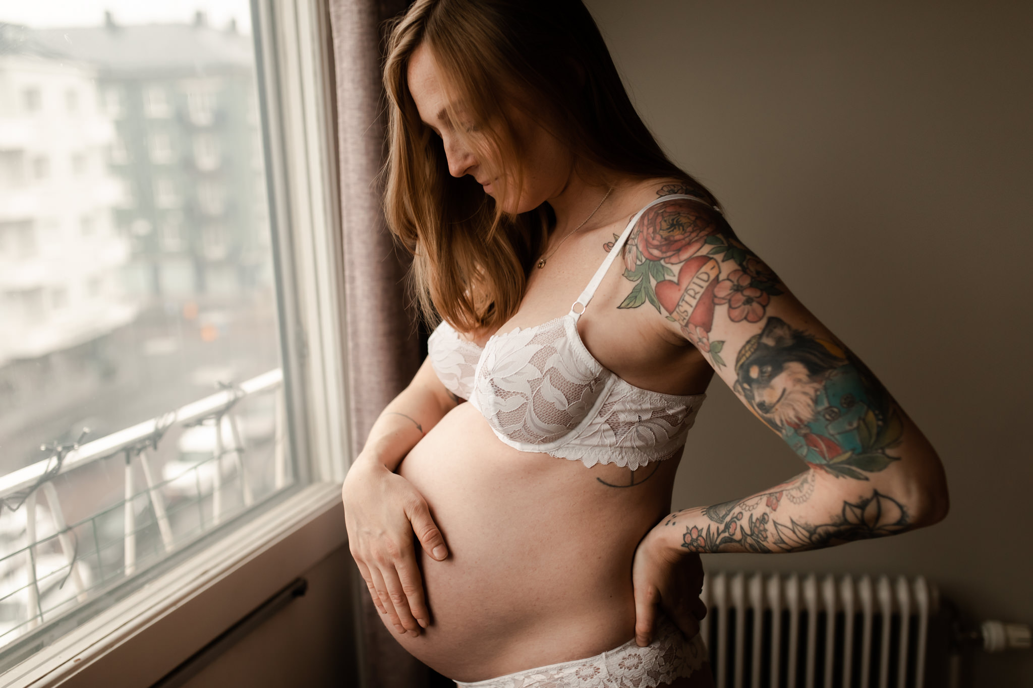 Gravid kvinna står framför ett fönster iklädd endast underkläder, och tittar på den gravida magen