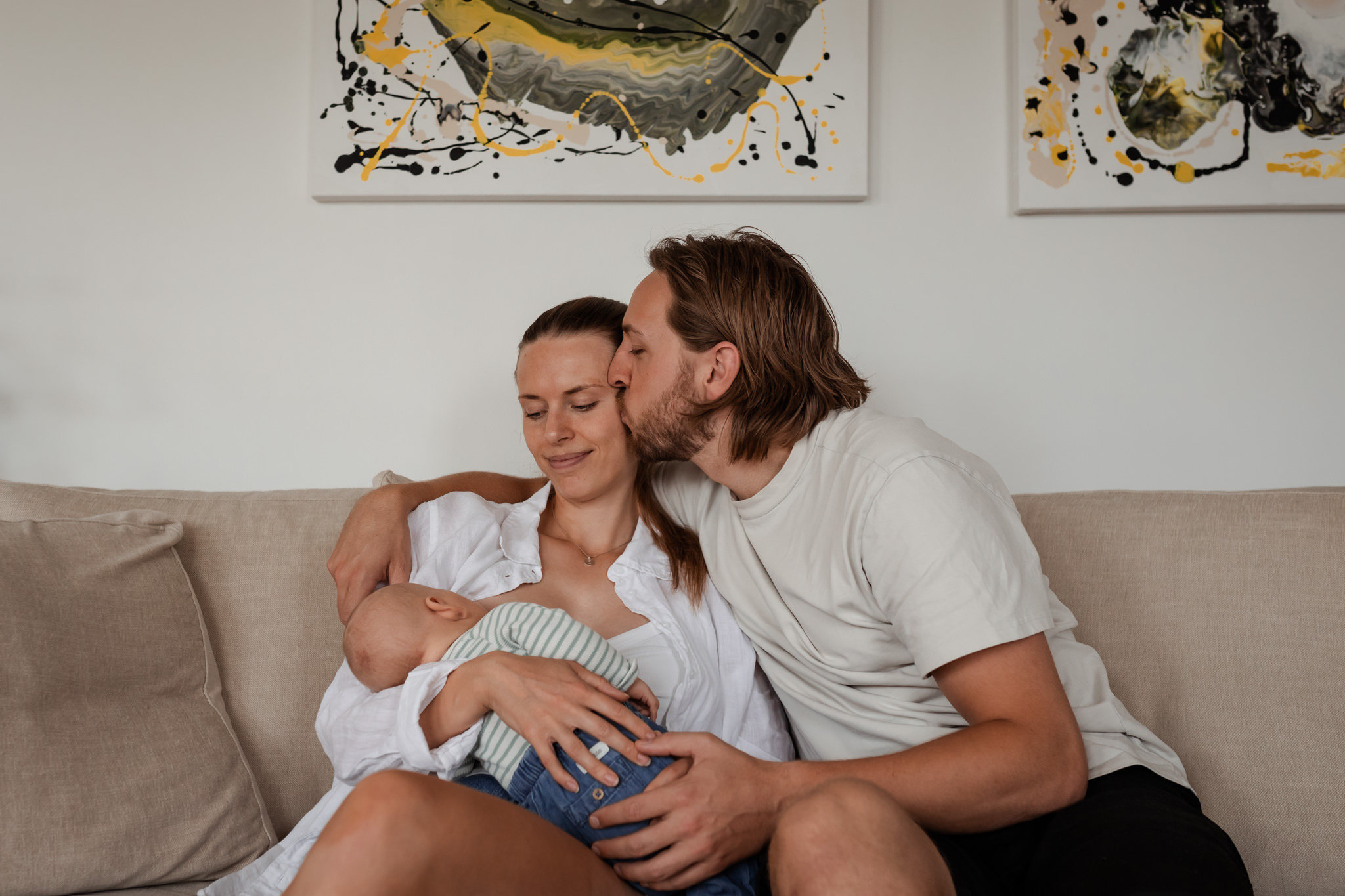Nyblivna föräldrar sitter i soffan i sitt hem med bebisen i famnen och pappan pussar på mammans tinning