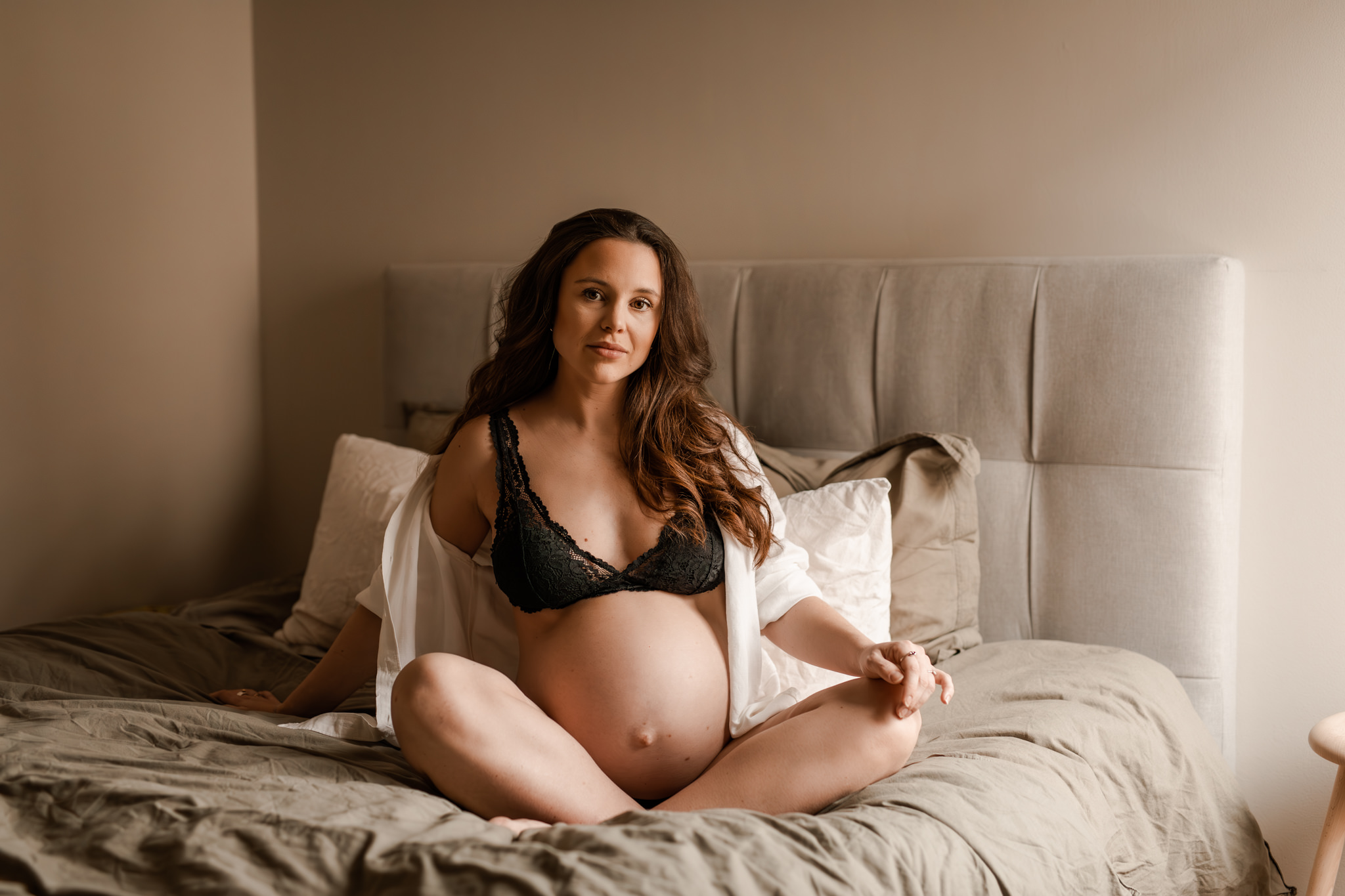 Gravid kvinna sitter på en säng iklädd bh, skjorta och jeans