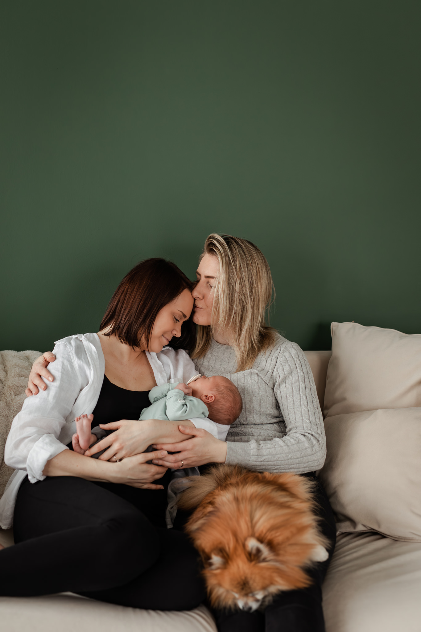 Samkönat kvinnligt par sitter i en soffa hållandes en nyfödd bebis, en liten hund i knäet och där den ena kvinnan pussar den andra på pannan