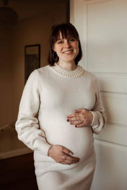 Gravid kvinna i page och lugg bär en vit stickad klänning och står och lutar sig mot en dörr och håller på magen på sin gravidfotografering i Småland