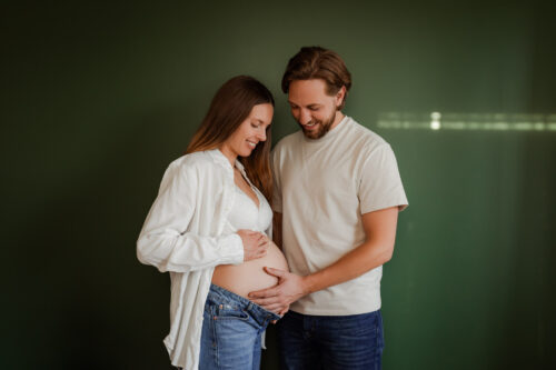 En gravid kvinna klädd i bh, jeans och vit skjorta står tillsammans med sin partner framför en grön vägg, hållandes händerna på den gravida magen.