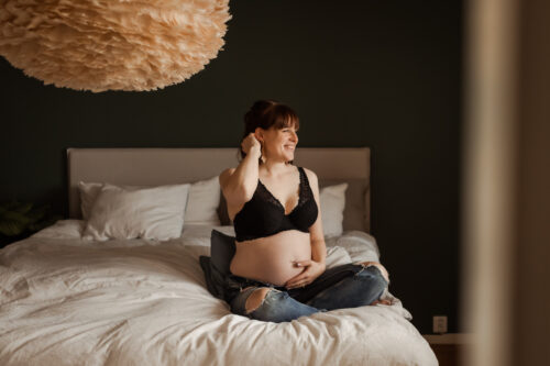 Gravid kvinna sitter i profil på en säng med bh och jeans och fixar med håret