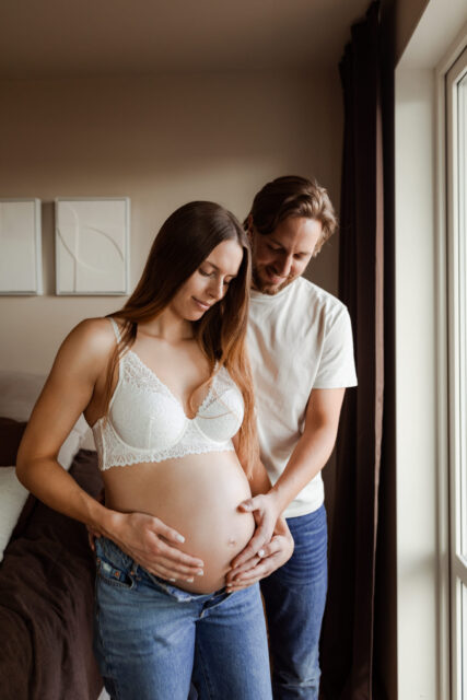 En gravid kvinna står med jeans och bh framför ett fönster medan hennes man håller en hand på hennes gravidmage under deras gravidfotografering i Småland