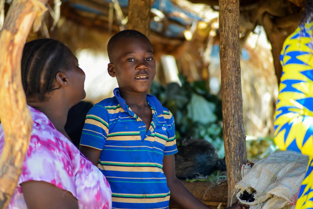 A boy named McLeans standing next to his mother and their produce stand at the market.