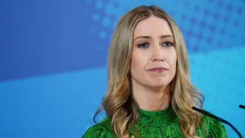 PA Media The head and shoulders of Laura Trott MP. She is wearing a green dress and stands against a textured blue background. She is framed to the right, with a microphone entering the shot in the bottom right hand corner.