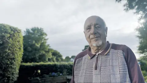 BBC/Hardcash Productions/Simon Rawles John Morris, with a white beard, stands in a garden and looks wryly at the camera.