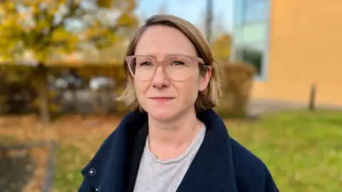 Steve Hubbard/BBC Saoirse Kerrigan is wearing a dark blue coat and a grey T-shirt. She has light pink coloured almost transparent glasses and has short bobbed mousey blonde hair. She is looking at the camera and is photographed outside.