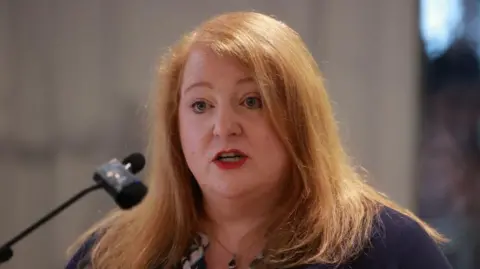 PA Media Long is standing in front of a microphone. She has long ginger hair and is wearing red lipstick. She has a navy top. 