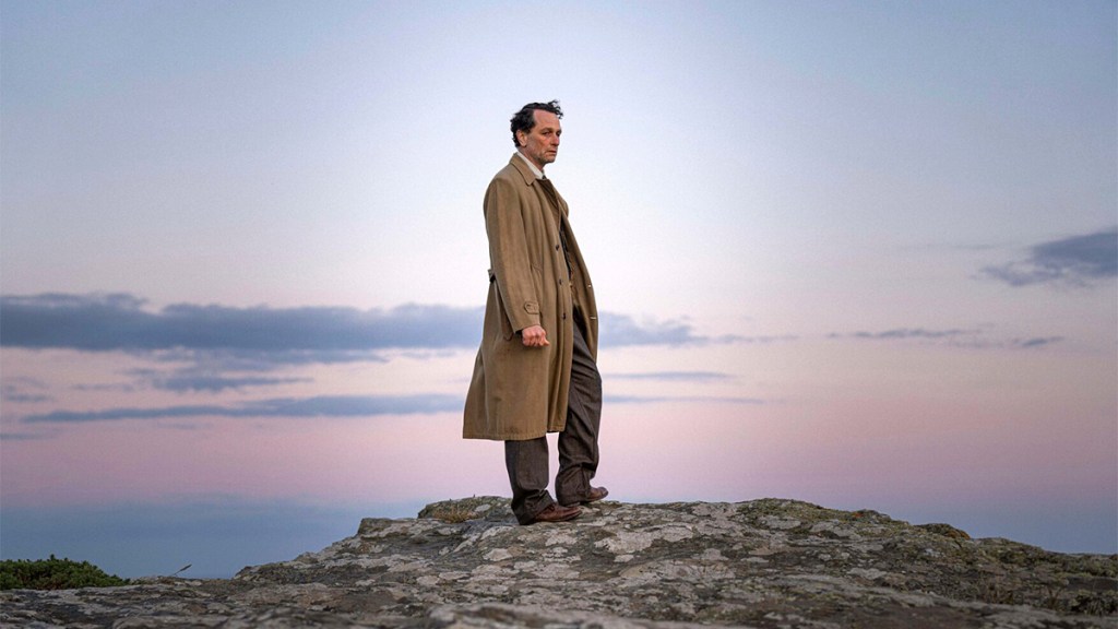 Matthew Rhys in a trenchcoat standing on a rocky outcrop