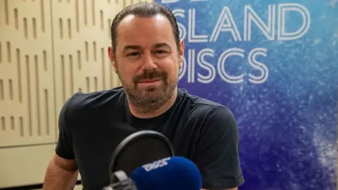 BBC Danny Dyer sits smiling in front of a Desert Island Discs backdrop.