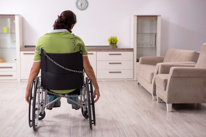 Mann i rullestol sitter alene i et rom med ryggen til. Ser trist ut.